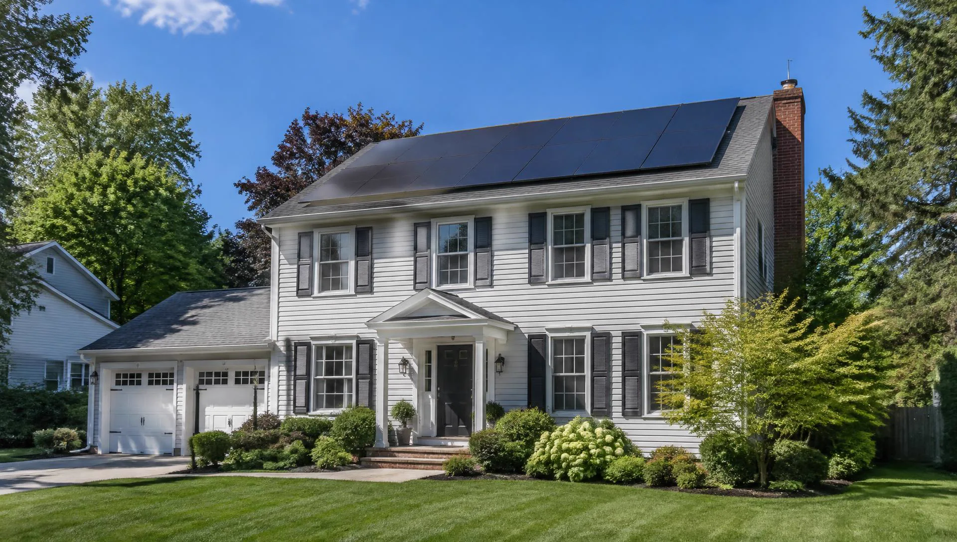 A suburban home with rooftop solar panels, a clear blue sky, and trees in the yard.